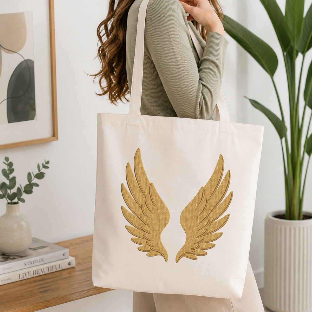 A person wearing a sage green ribbed sweater carries a cream canvas tote bag with a large gold wing graphic on the front, set in a bright, modern room with a wooden table, framed art, and a potted plant.