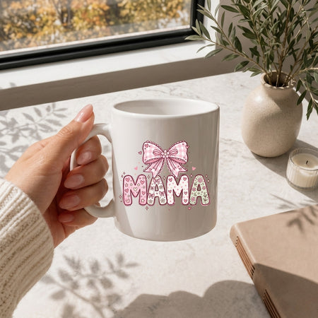 Hand holding a white ceramic coffee mug printed with a pastel pink patterned "MAMA" coquette bow design on a bright table.