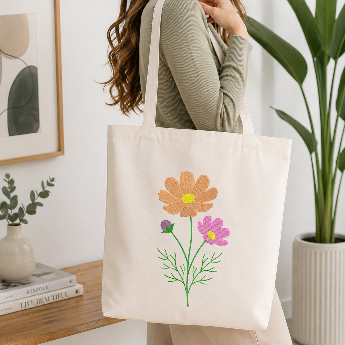 A woman wearing a sage green ribbed long-sleeve top carries a cream canvas tote bag decorated with embroidered flowers in orange, pink, purple, and green. The setting is a bright, modern room with a wooden table, books, a small vase, and a potted plant.