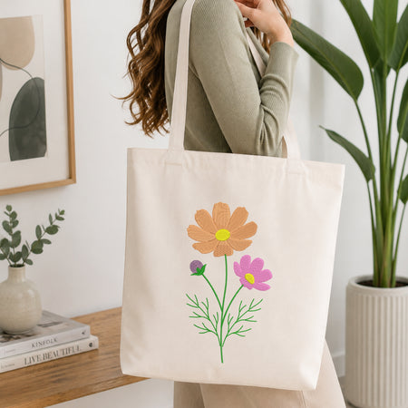 A woman wearing a sage green ribbed long-sleeve top carries a cream canvas tote bag decorated with embroidered flowers in orange, pink, purple, and green. The setting is a bright, modern room with a wooden table, books, a small vase, and a potted plant.