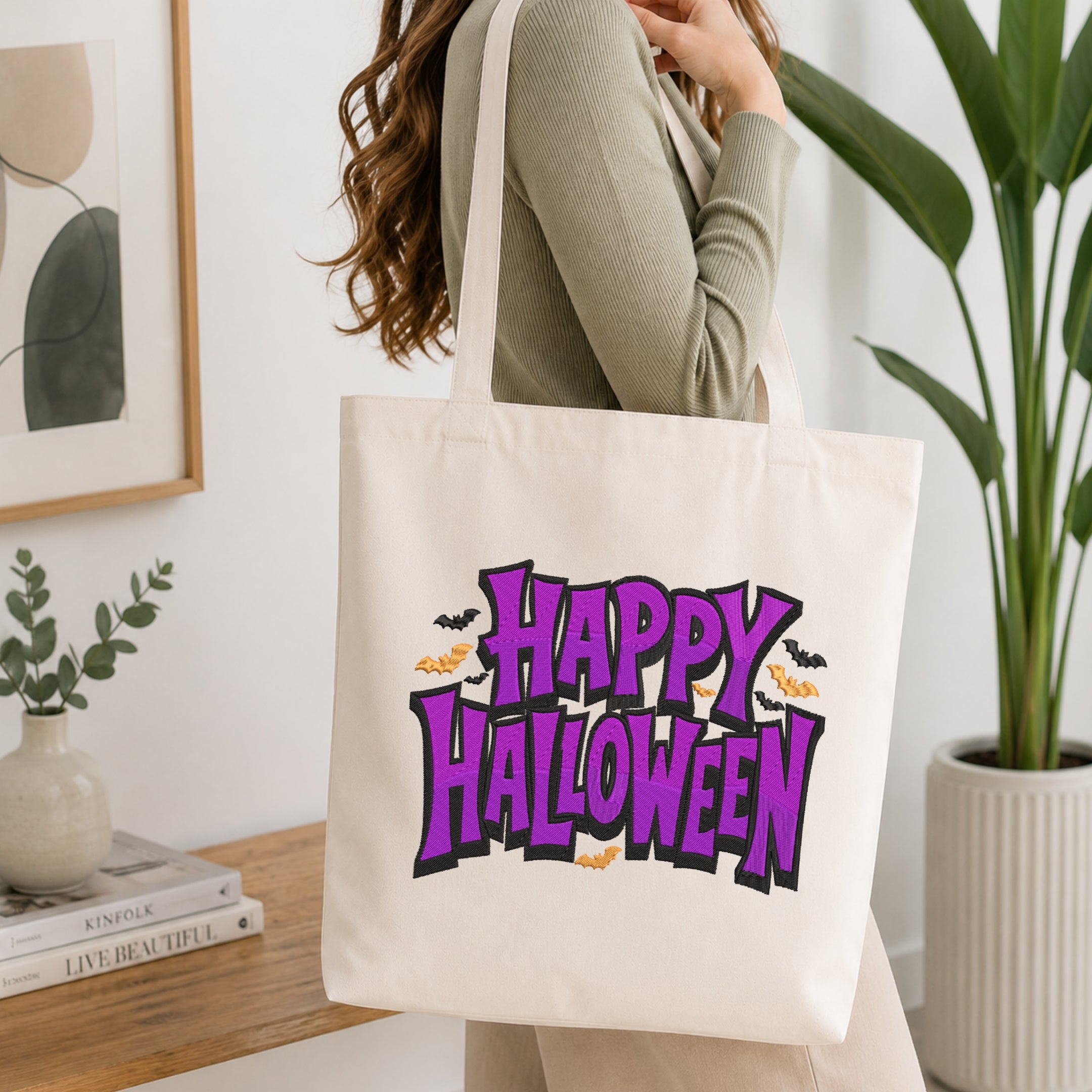A woman wearing a sage green ribbed long-sleeve top carries a cream canvas tote bag with a colorful 'Happy Halloween' graphic in purple, orange, and black. The photo is taken in a bright, modern room with a wooden console table, a potted green plant, and a framed abstract print.