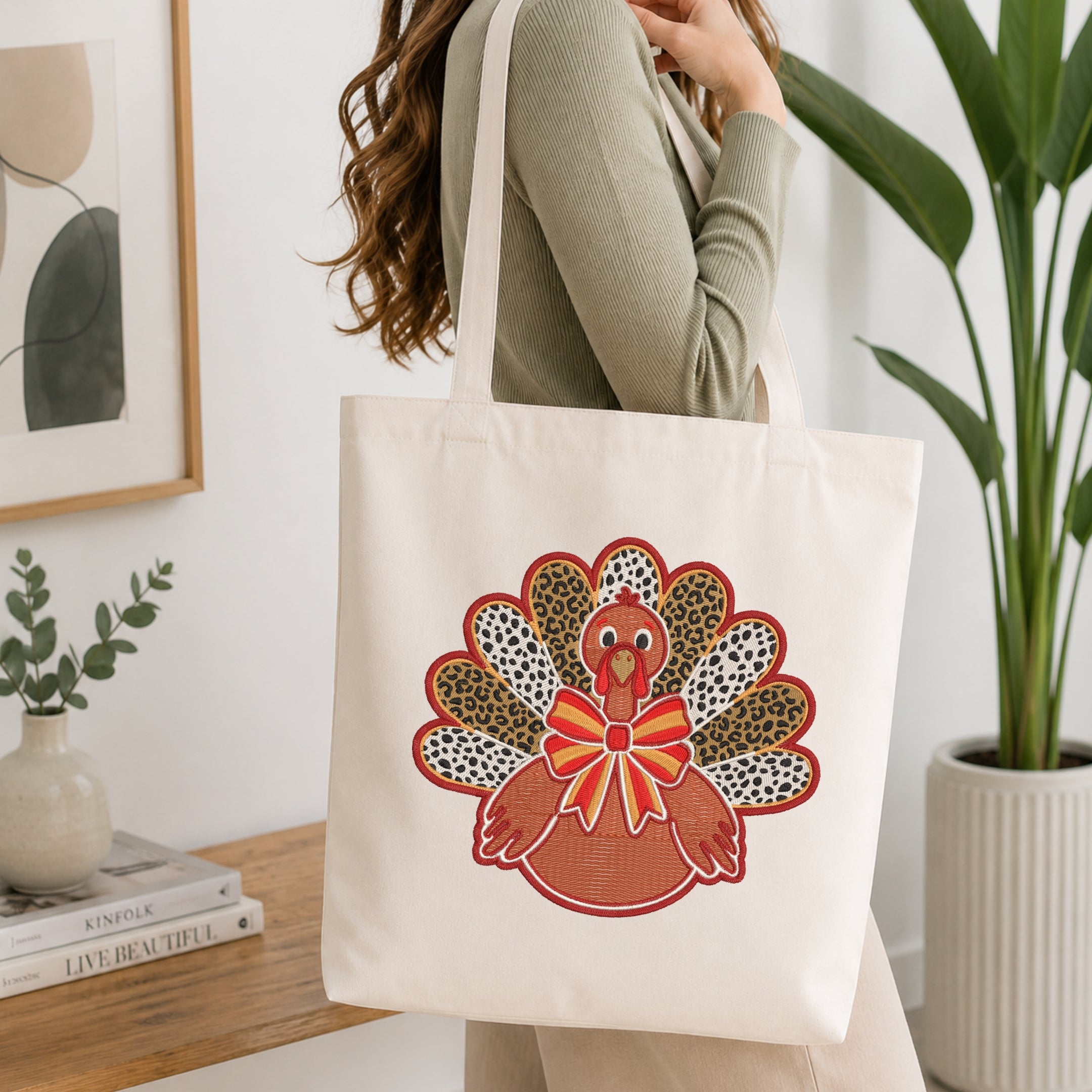 A woman wearing a sage green ribbed sweater carries a cream canvas tote bag with a colorful embroidered turkey design featuring leopard print feathers and a red bow, set in a bright, modern room with a wooden table and potted plants.