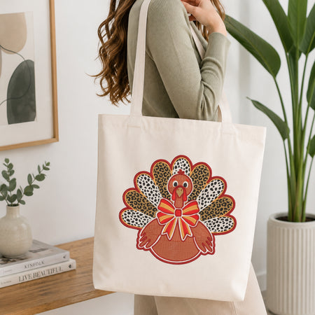 A woman wearing a sage green ribbed sweater carries a cream canvas tote bag with a colorful embroidered turkey design featuring leopard print feathers and a red bow, set in a bright, modern room with a wooden table and potted plants.