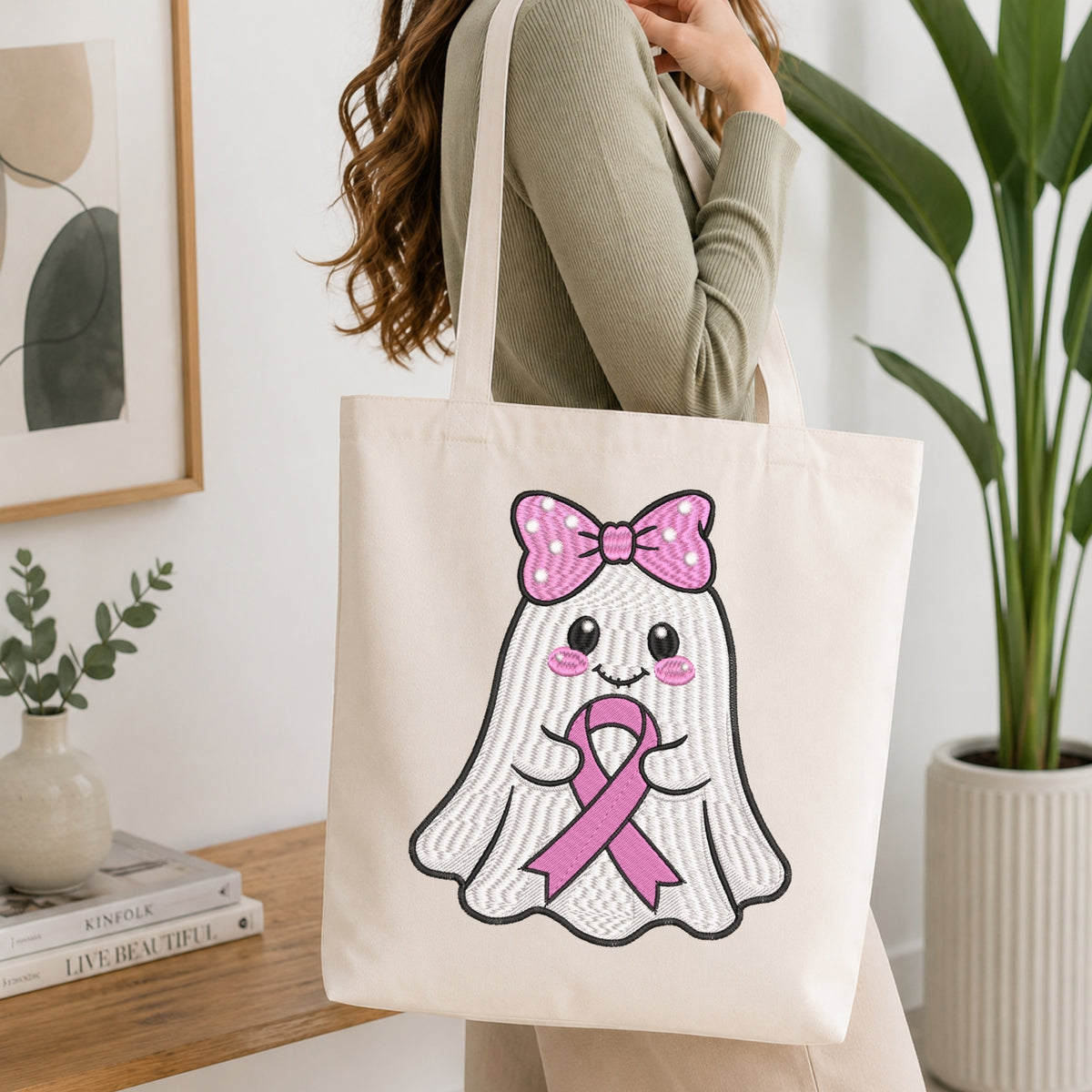 A woman wearing a sage green ribbed long-sleeve top carries a cream canvas tote bag with a cute embroidered ghost graphic wearing a pink bow and pink ribbon.