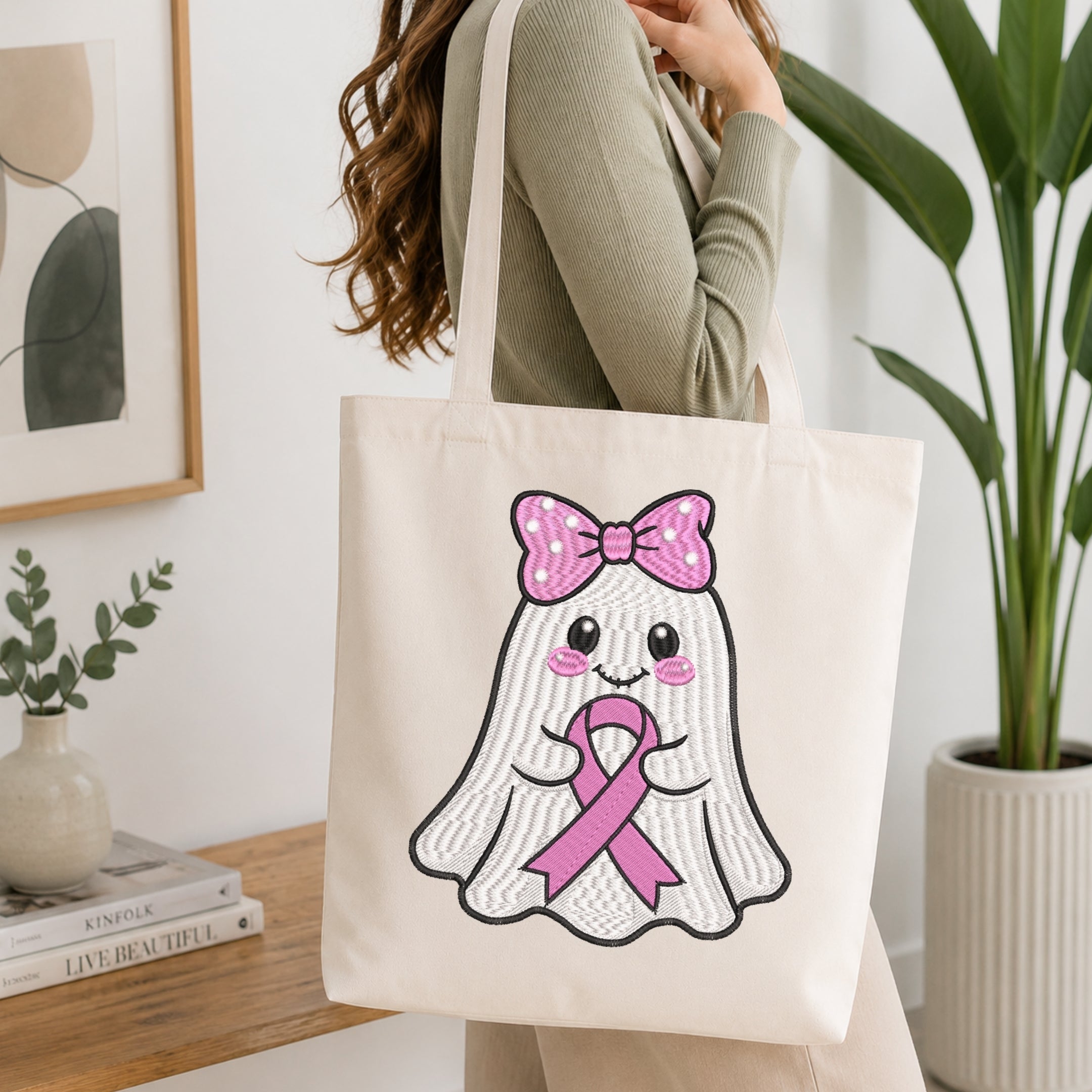 A woman wearing a sage green ribbed long-sleeve top carries a cream canvas tote bag with a cute embroidered ghost graphic wearing a pink bow and pink ribbon.