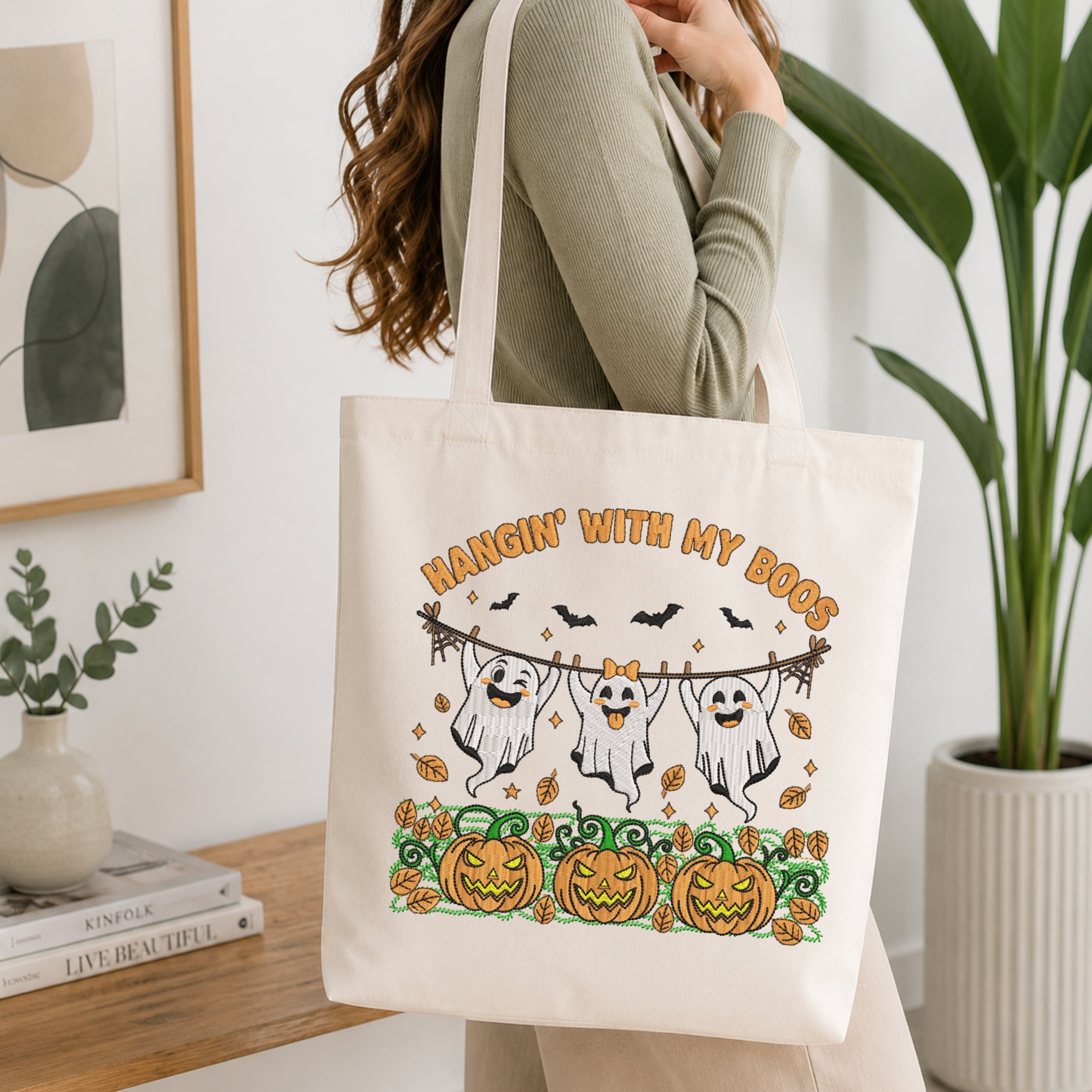 A woman wearing a sage green ribbed long-sleeve top carries a cream canvas tote bag with a Halloween graphic that reads 'HANGIN' WITH MY DOGS' above three cartoon ghosts and pumpkins. The scene is set in a bright, modern room with a wooden table, books, a plant, and framed wall art.