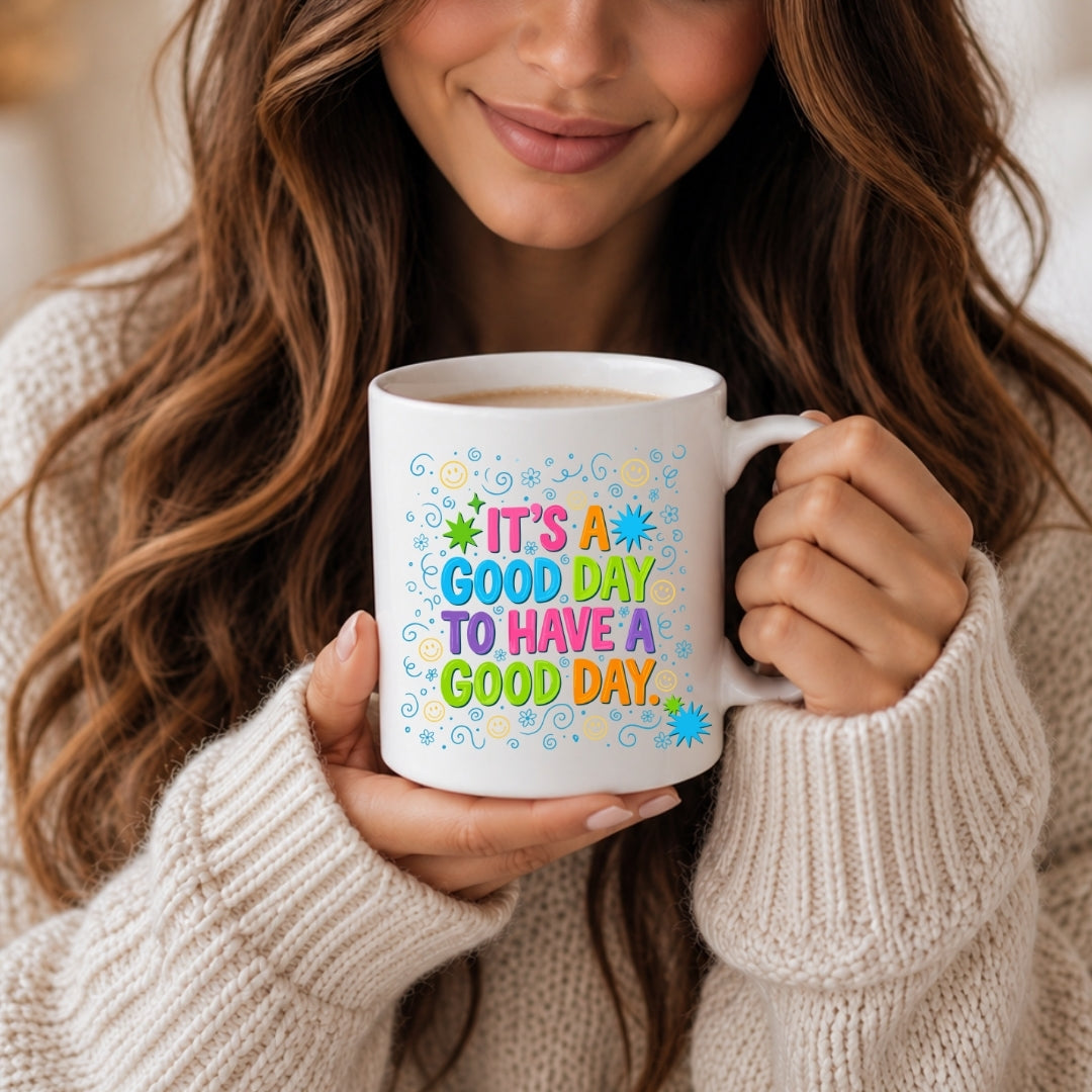 A woman in a cream knit sweater holds a white ceramic mug with colorful text that reads 'IT'S A GOOD DAY TO HAVE A GOOD DAY.' The mug has a playful multicolor design with stars, dots, and wavy lines.