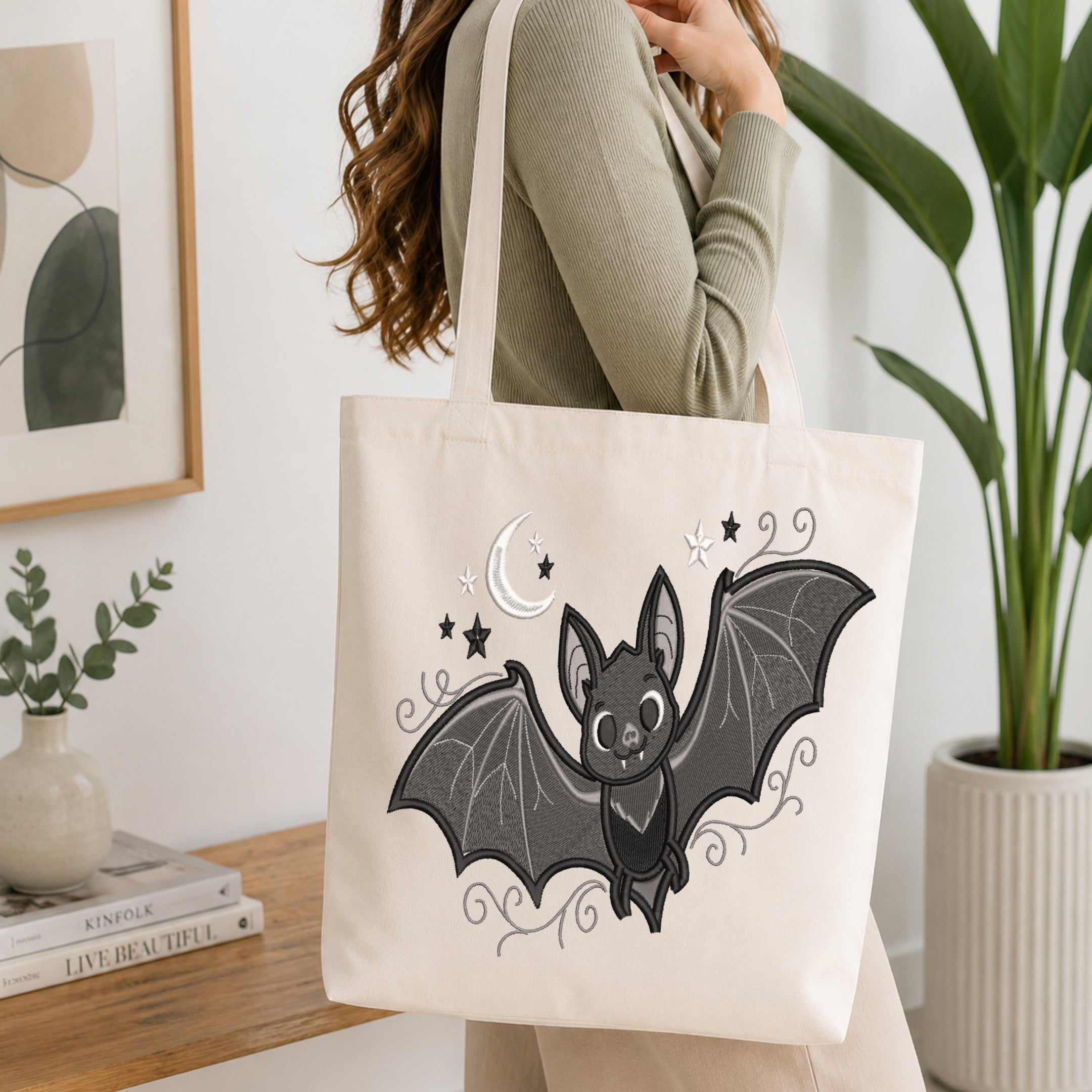 An off-white canvas tote bag with a black and gray bat illustration, carried by a woman in a ribbed sage green top in a bright, modern room.
