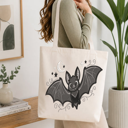 An off-white canvas tote bag with a black and gray bat illustration, carried by a woman in a ribbed sage green top in a bright, modern room.