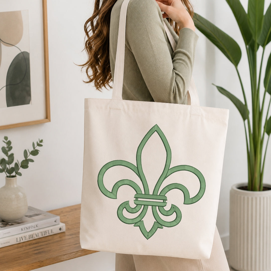 An off-white canvas tote bag with a green fleur-de-lis design, carried over the shoulder of a person wearing a sage green ribbed sweater in a bright, modern room with a wooden table and potted plants.
