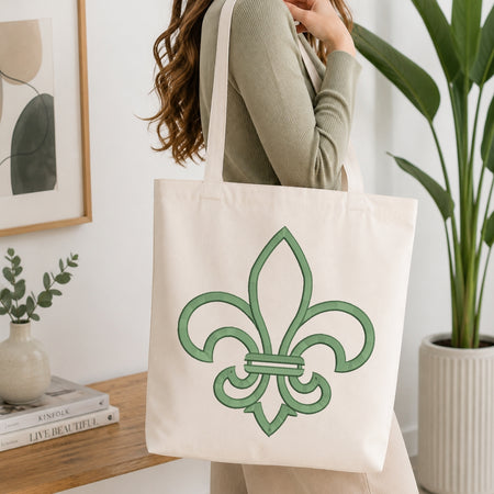 An off-white canvas tote bag with a green fleur-de-lis design, carried over the shoulder of a person wearing a sage green ribbed sweater in a bright, modern room with a wooden table and potted plants.