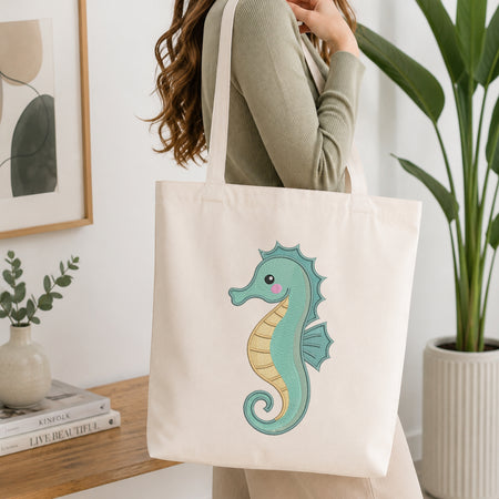 A woman wearing a sage green ribbed long-sleeve top carries a cream canvas tote bag featuring a cute, colorful embroidered seahorse. The setting is a bright, modern room with a wooden side table, a potted plant, and framed wall art.