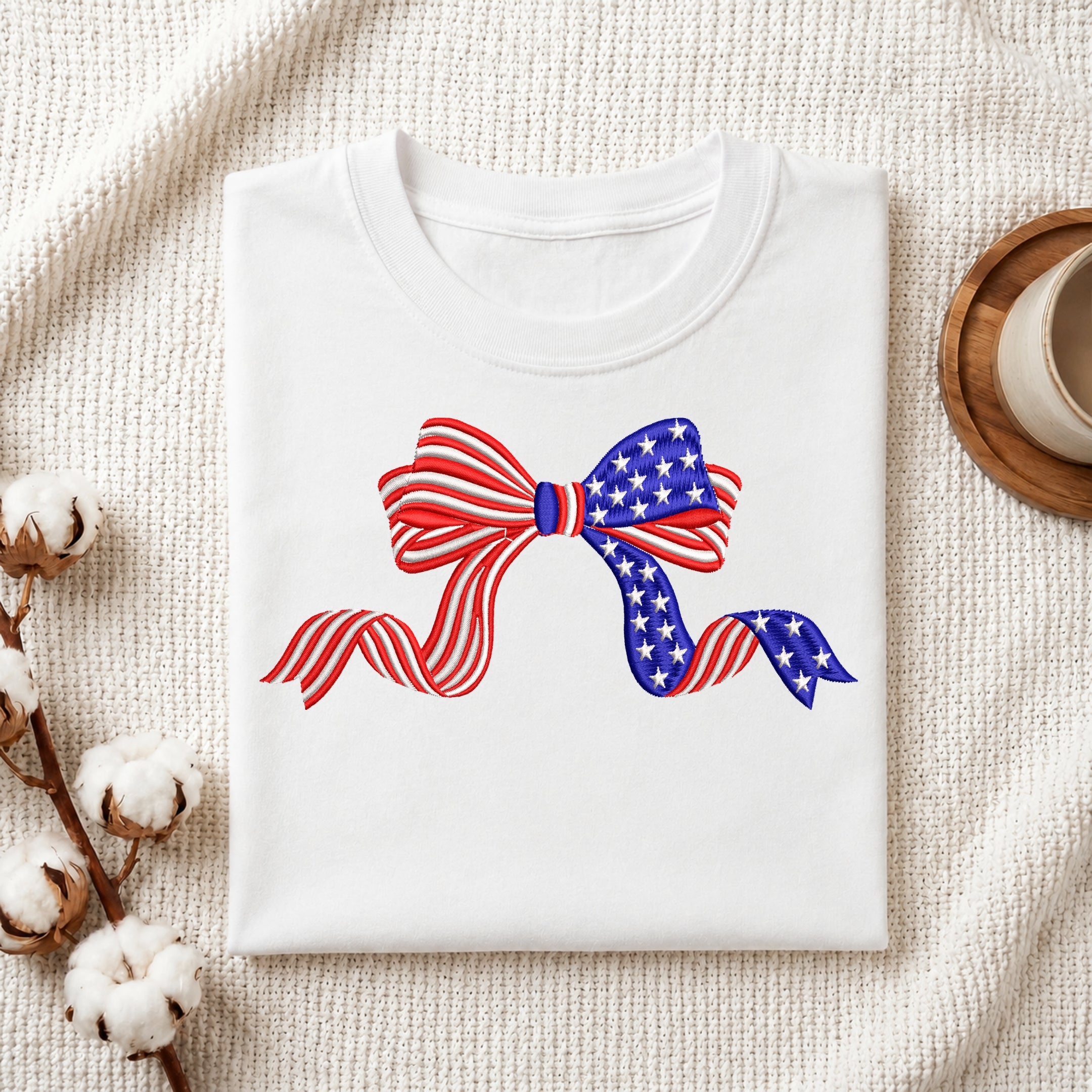 A folded white crewneck t-shirt featuring a graphic of a ribbon bow styled with the American flag pattern (red and white stripes on one side, white stars on blue on the other), displayed on a textured cream fabric background with decorative cotton stems and a wooden cup.