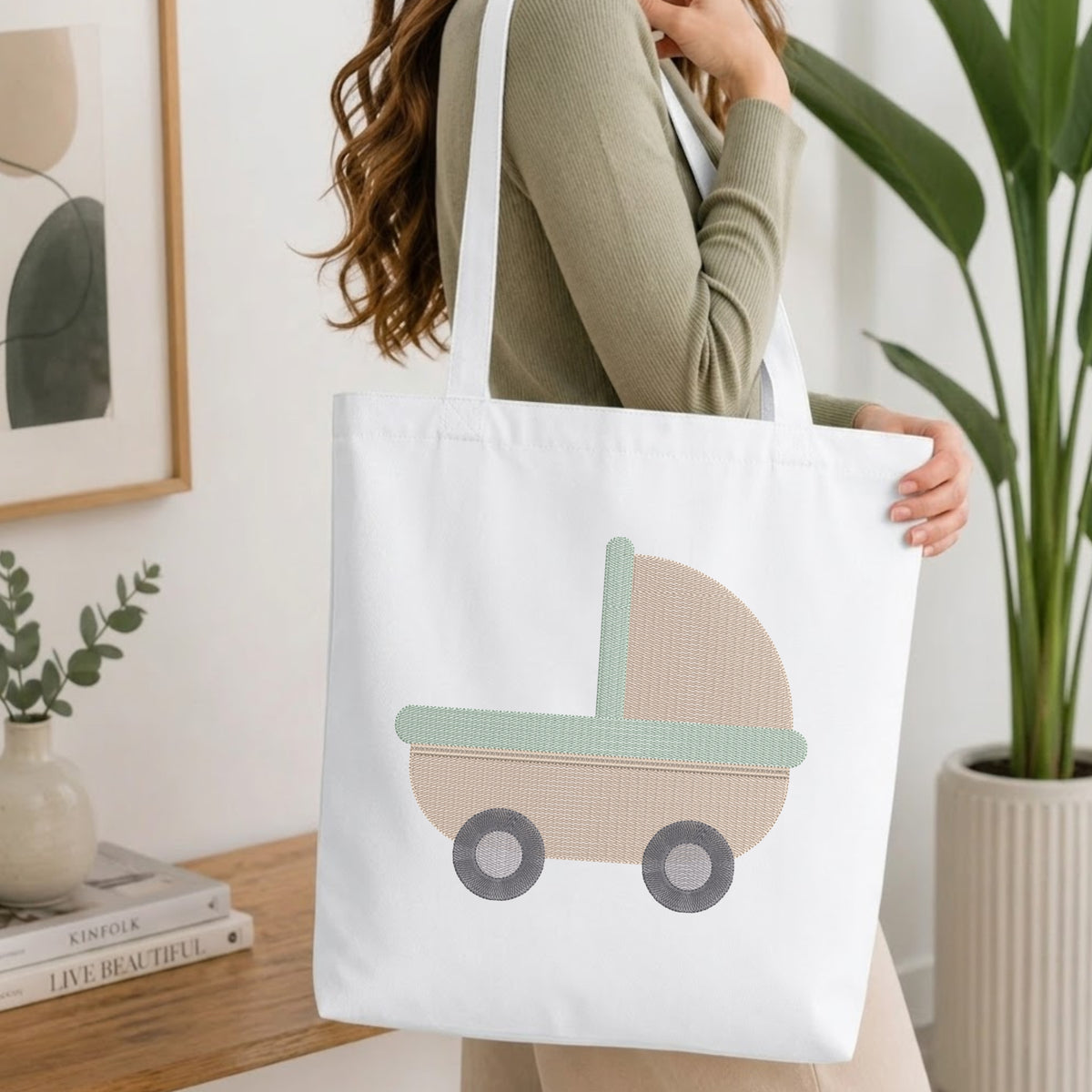 A woman in a sage green ribbed sweater carries a white canvas tote bag featuring a stylized baby stroller design in beige, mint green, and gray. The setting is a bright, modern room with a wooden table, books, a plant, and framed wall art.