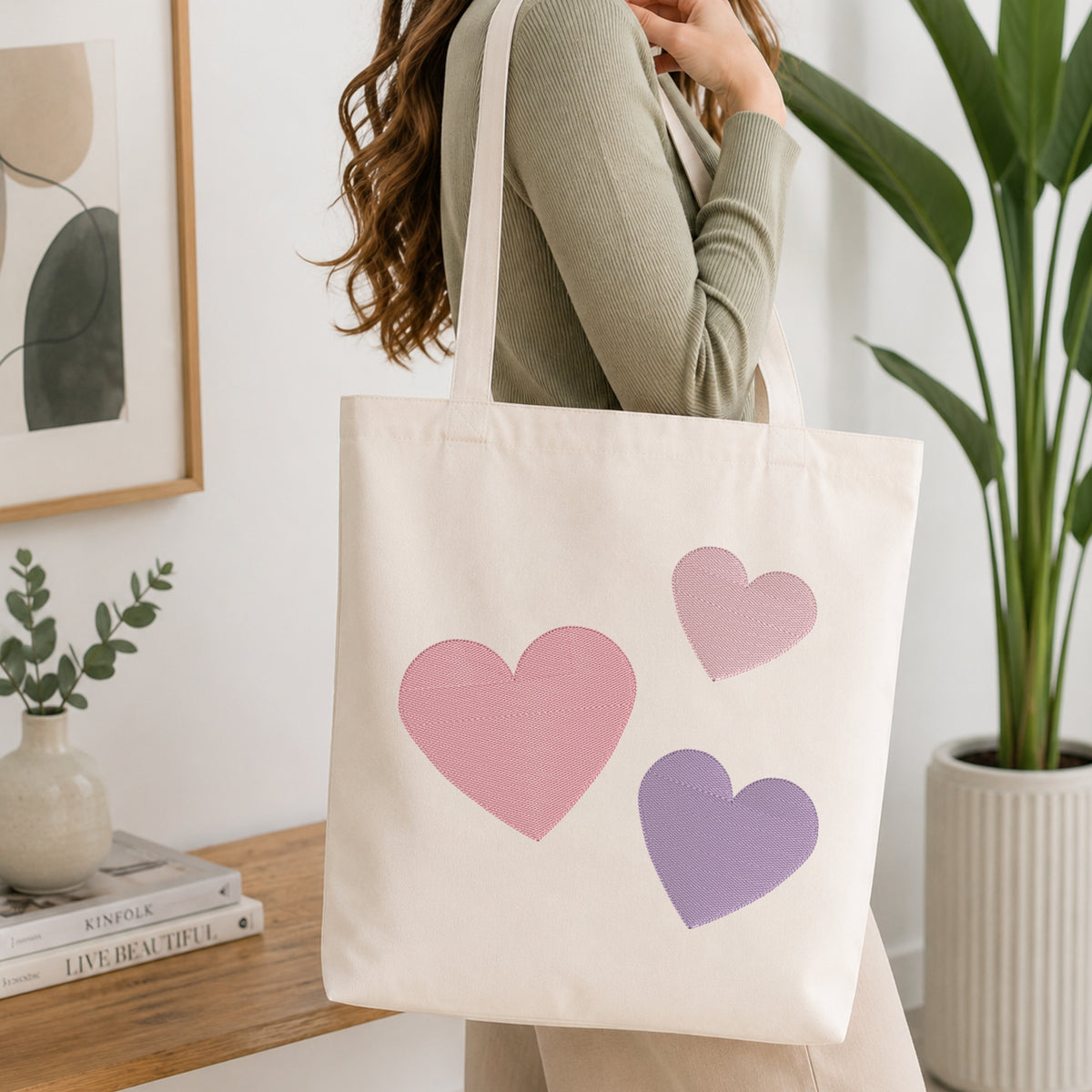 A person wearing a sage green ribbed long-sleeve top carries a cream canvas tote bag with three embroidered hearts in pink and purple, set against a bright, modern room background with a wooden table and potted plants.
