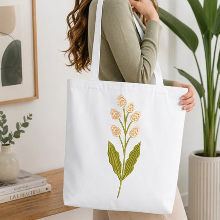 A woman wearing a sage green ribbed long-sleeve top carries a white canvas tote bag with an embroidered floral design of peach flowers and green leaves, set in a bright, modern room with a wooden table and potted plants.