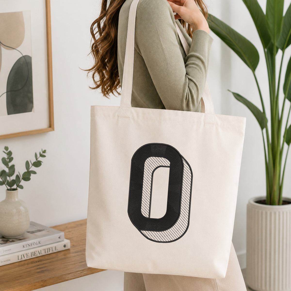 A woman wearing a sage green ribbed long-sleeve top carries a cream canvas tote bag with a large black graphic letter 'O' on the front, set in a bright, modern room with a wooden console table, framed art, and a potted plant.