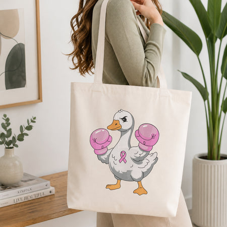 A woman wearing a sage green ribbed long-sleeve top carries a cream canvas tote bag. The bag features a cartoon illustration of a white duck with orange beak and feet, wearing pink boxing gloves and a pink ribbon on its chest.