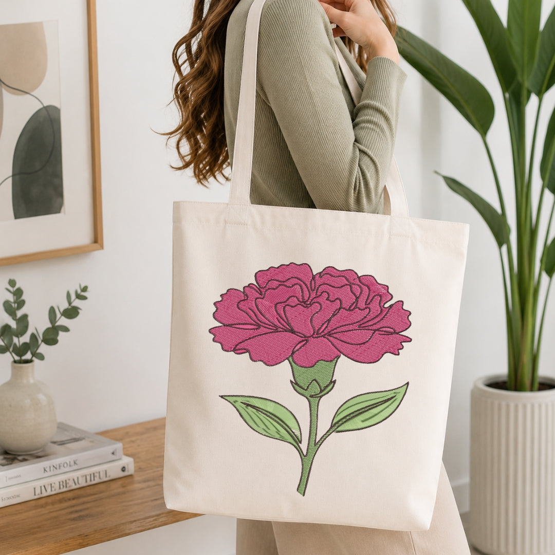 A woman wearing a sage green ribbed sweater carries a cream canvas tote bag with a large pink flower and green leaves printed on the front, set in a bright, modern room with a wooden table and potted plants.