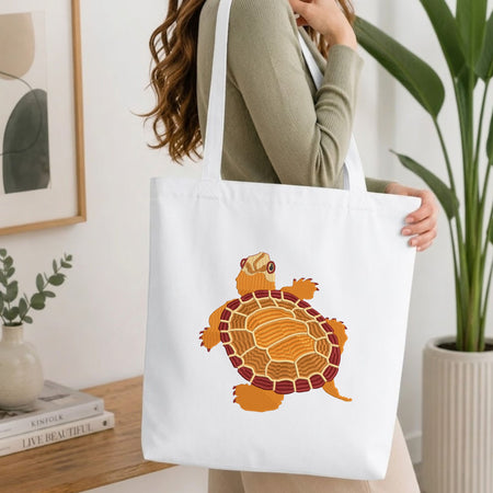 Woman carrying a white natural canvas tote bag featuring an embroidered turtle design in a bright, modern interior setting   