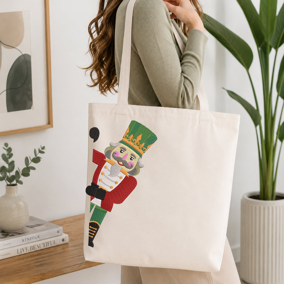 A person wearing a green long-sleeve top and carrying a natural canvas tote bag printed with the peeking nutcracker Christmas machine embroidery graphic.