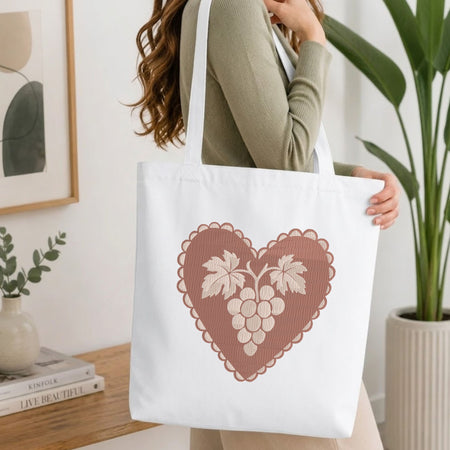 A woman wearing a sage green ribbed long-sleeve top carries a white canvas tote bag with a large dusty rose and cream heart-shaped grapevine graphic on the front, set in a bright, modern room with a wooden table and potted plants.