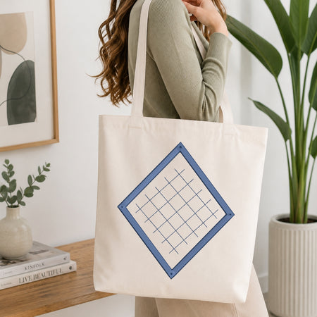 A woman wearing a sage green ribbed sweater carries a cream canvas tote bag with a blue geometric grid design on the front, set in a bright, modern room with a wooden table, books, and a potted plant.