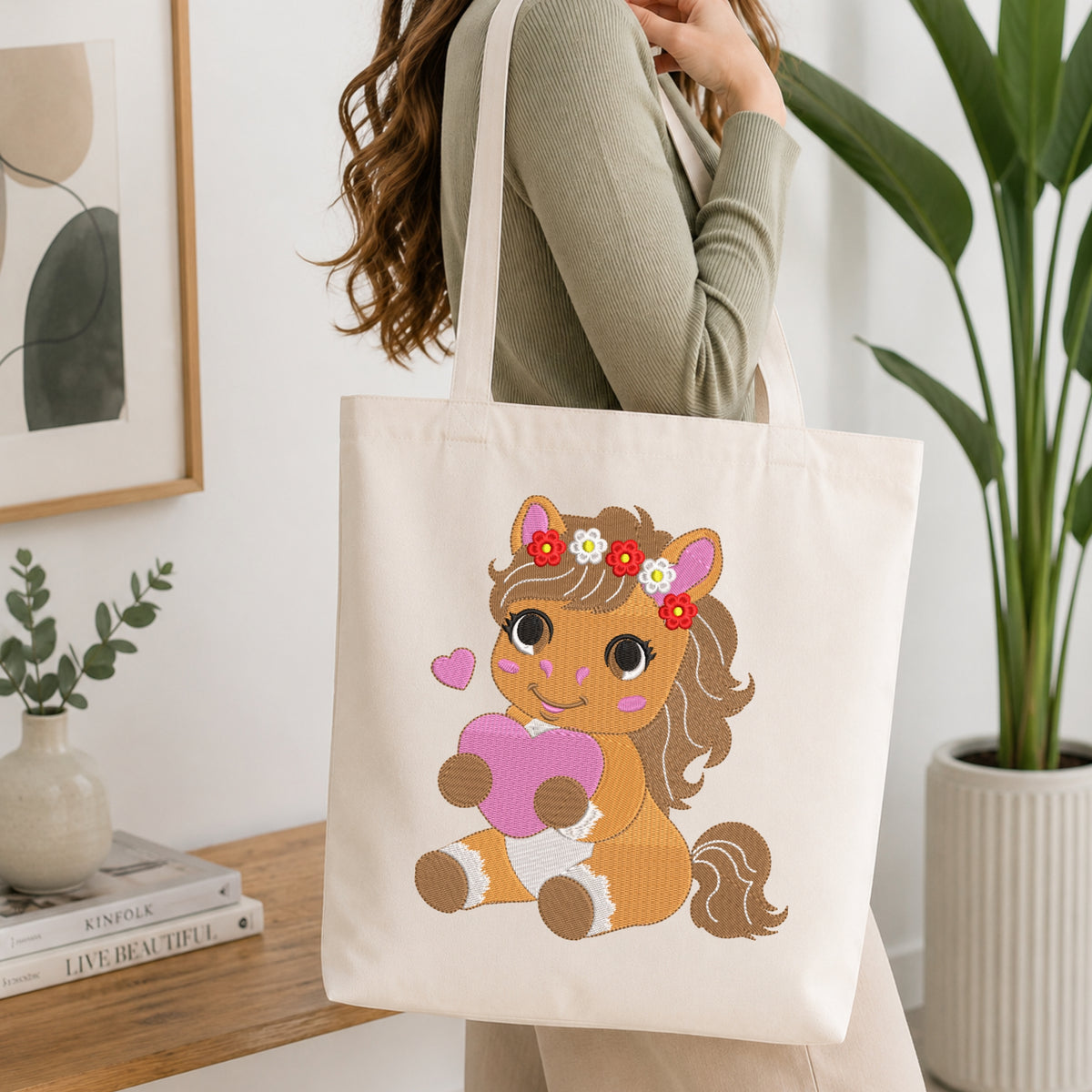 A person wearing a sage green ribbed long-sleeve top carries a cream canvas tote bag with a cartoon horse graphic. The horse is brown with a pink heart, red and white flowers in its mane, and pink cheeks.