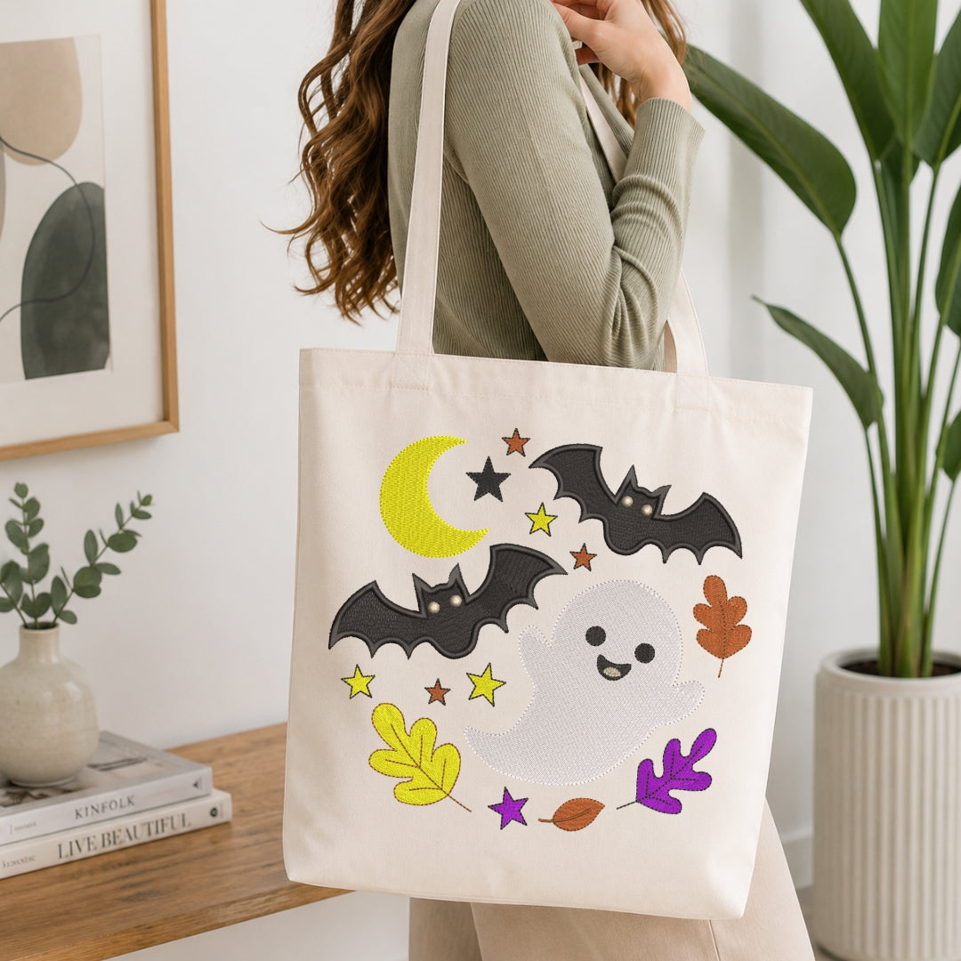 A woman wearing a sage green ribbed sweater carries a cream canvas tote bag with a Halloween design. The design features two black bats, a smiling white ghost, a yellow crescent moon, and colorful autumn leaves (yellow, purple, orange, brown) on a white background.