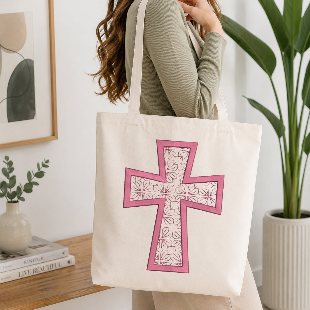 A person wearing a ribbed sage green top carries a cream canvas tote bag with a large pink cross design featuring a floral pattern, set in a bright, modern room with a wooden table and potted plants.
