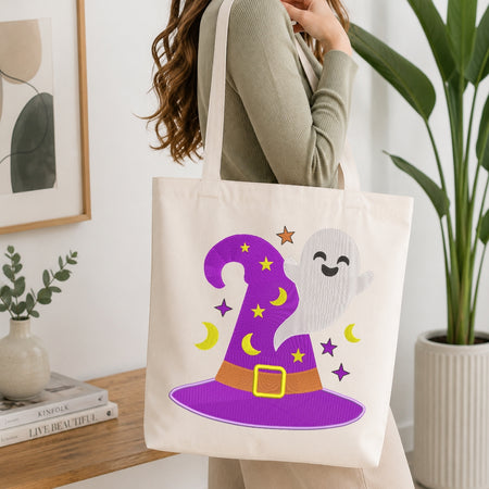 A person wearing a sage green ribbed long-sleeve top carries a cream canvas tote bag featuring a purple witch hat and a smiling white ghost with yellow crescent moons and stars.
