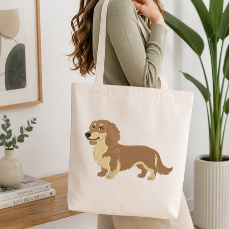 A person holding a natural canvas tote bag printed with an embroidered brown dachshund dog illustration, set against a bright interior room background.