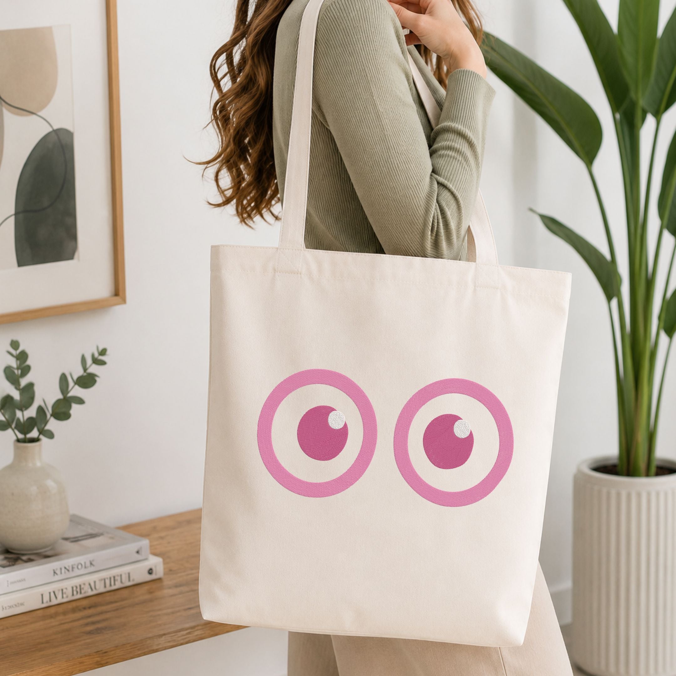 A woman wearing a sage green ribbed sweater carries a cream canvas tote bag with a pink graphic of two cartoon eyes. The setting is a bright, modern room with a wooden table, books, a small plant, and a large green potted plant in the background.