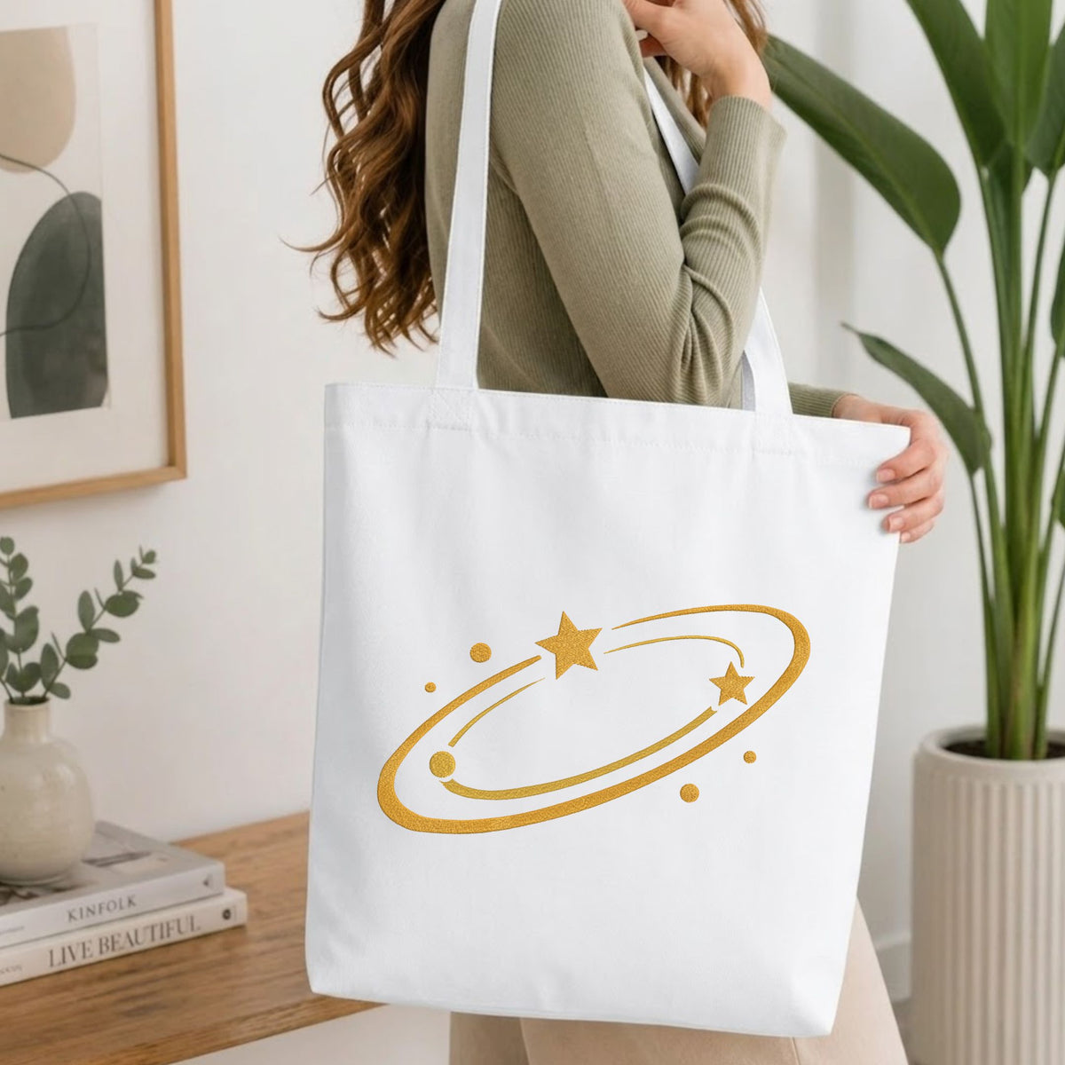 Woman carrying a white natural canvas tote bag featuring an embroidered golden swirling star design in a bright, modern interior setting