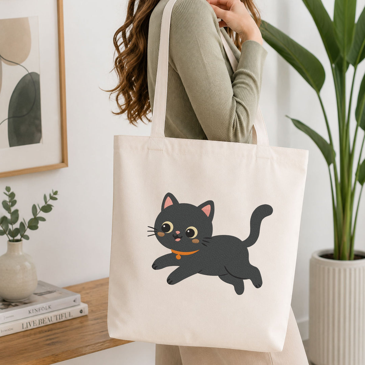 Woman carrying a white natural canvas tote bag featuring a cute playful black cat machine embroidery design in a bright, modern interior setting