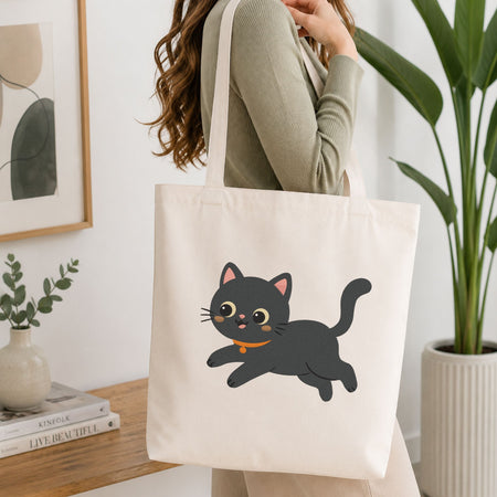 Woman carrying a white natural canvas tote bag featuring a cute playful black cat machine embroidery design in a bright, modern interior setting