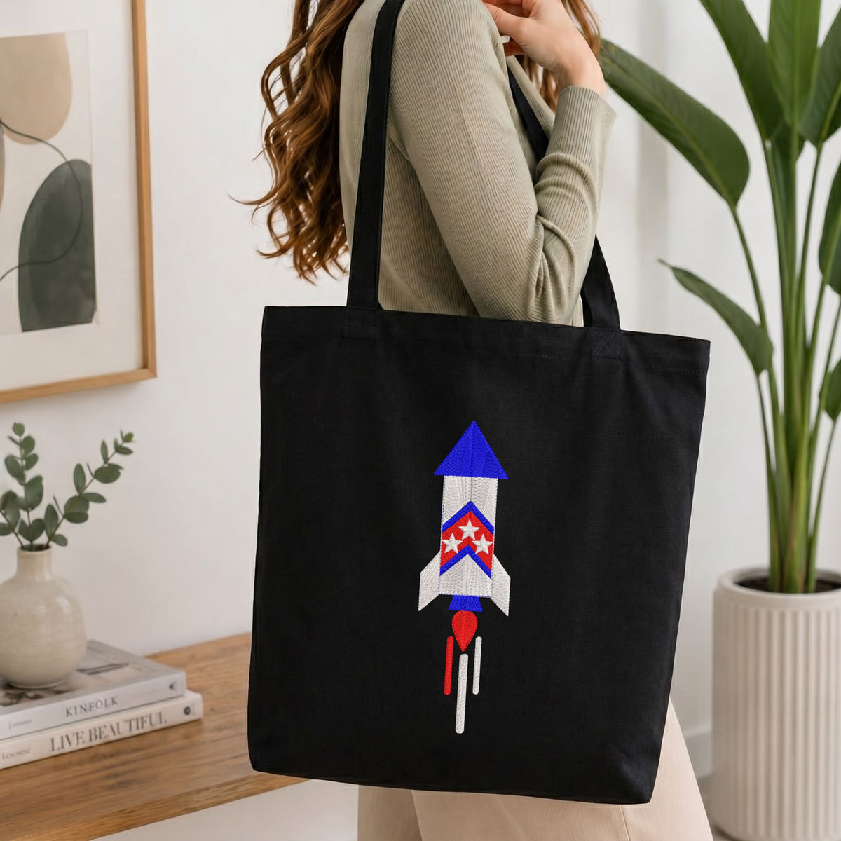 Woman carrying a black natural canvas tote bag featuring an embroidered patriotic space rocket design in a bright, modern interior setting.