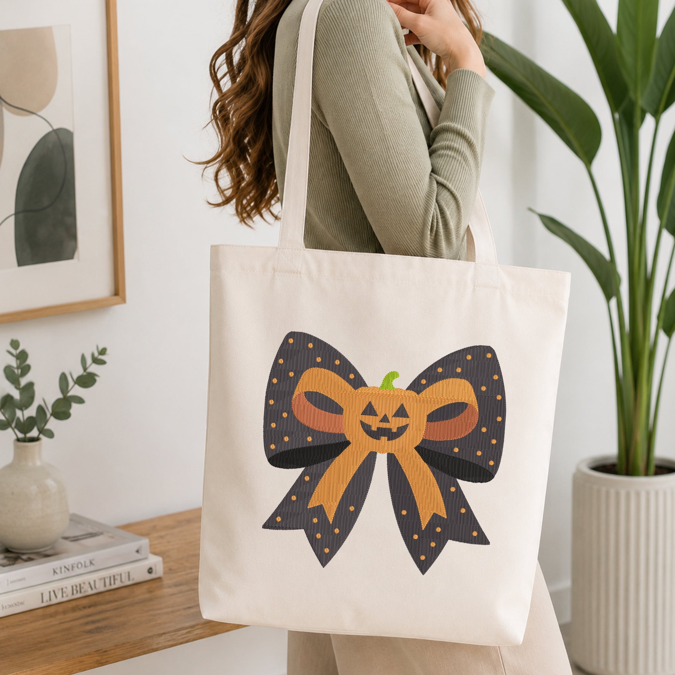 A person wearing a green long-sleeve top and carrying a natural canvas tote bag printed with the Halloween pumpkin bow machine embroidery graphic