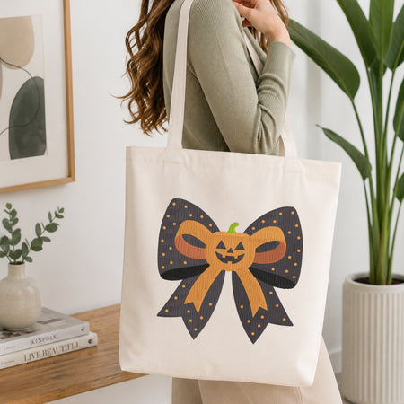 A person wearing a green long-sleeve top and carrying a natural canvas tote bag printed with the Halloween pumpkin bow machine embroidery graphic