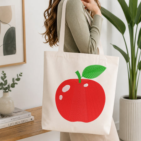 A cream canvas tote bag with a large red apple and green leaf graphic, carried by a woman in a ribbed sage green top in a bright, modern room.