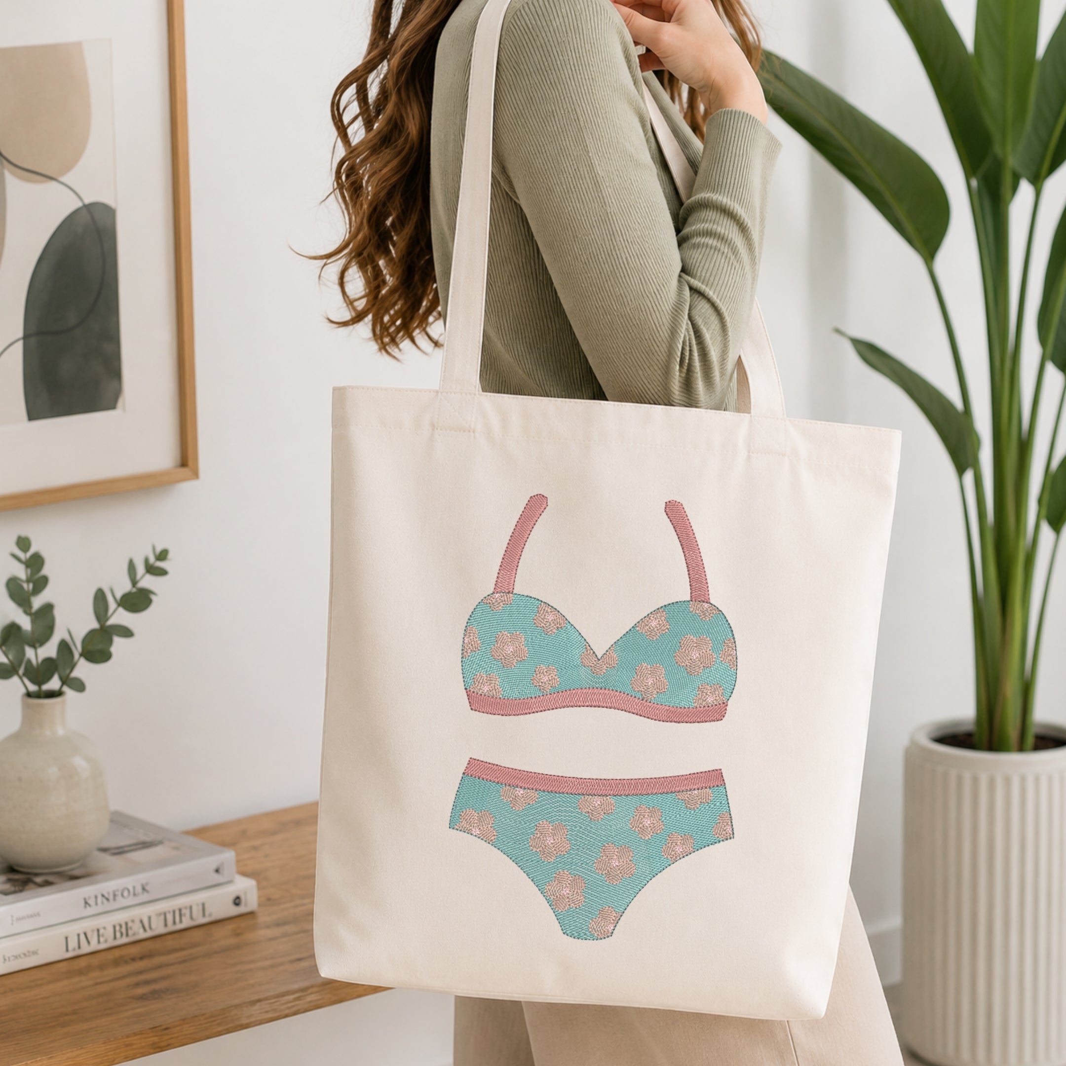 A woman wearing a sage green ribbed long-sleeve top carries a cream canvas tote bag. The bag features a printed graphic of a two-piece bikini in a light teal floral pattern with pink straps and trim.