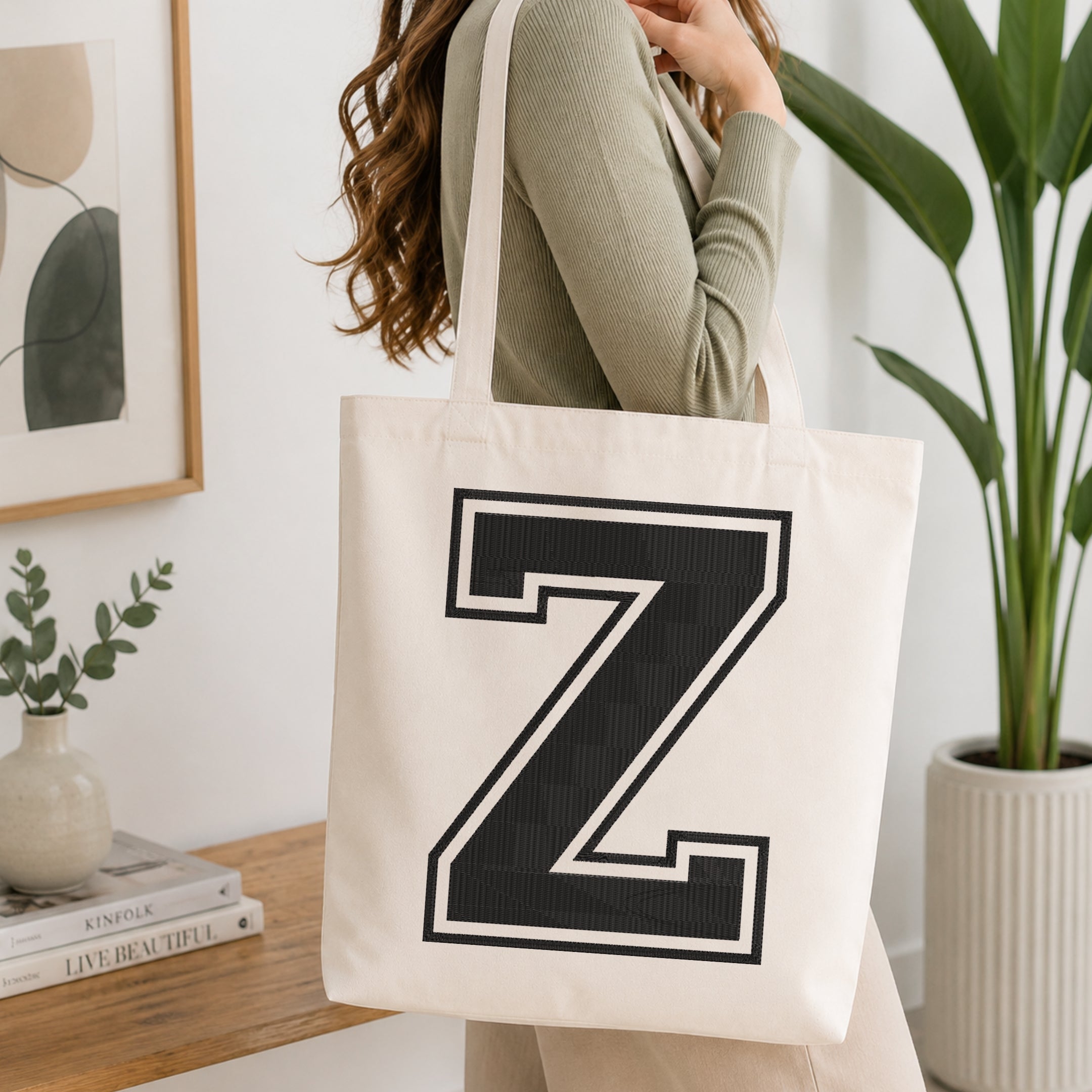 A woman wearing a sage green ribbed long-sleeve top carries a cream canvas tote bag with a large black collegiate-style letter 'Z' outlined in white on the front. The scene is set in a bright, modern room with a wooden console table, a framed print, a small vase with greenery, and a potted plant.