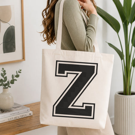 A woman wearing a sage green ribbed long-sleeve top carries a cream canvas tote bag with a large black collegiate-style letter 'Z' outlined in white on the front. The scene is set in a bright, modern room with a wooden console table, a framed print, a small vase with greenery, and a potted plant.
