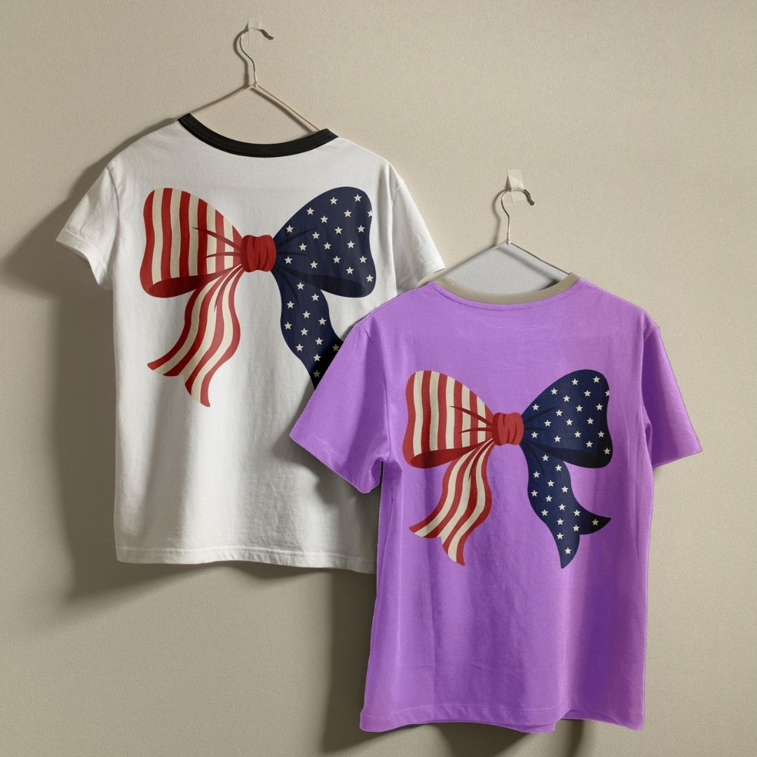 A white ringer t-shirt with a black collar and a solid purple t-shirt hanging on a wall, showcasing the custom patriotic bow design asset print.