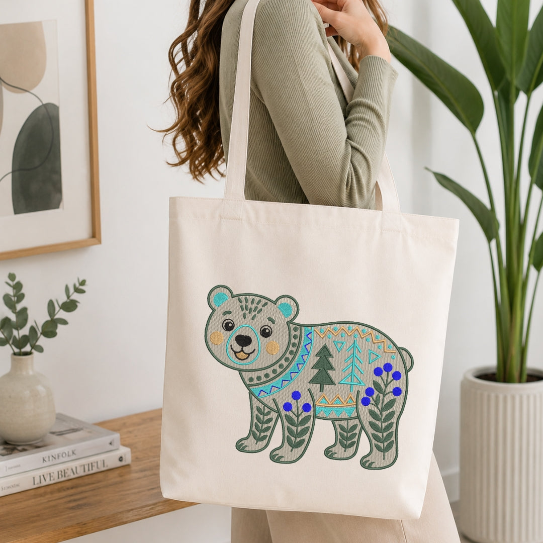 A person wearing a sage green ribbed sweater carries a cream canvas tote bag with a colorful embroidered polar bear design featuring blue flowers and green trees, set in a bright, modern room with a wooden table and potted plants.