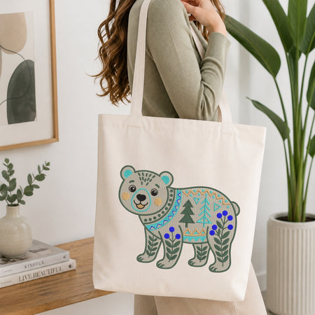 A person wearing a sage green ribbed sweater carries a cream canvas tote bag with a colorful embroidered polar bear design featuring blue flowers and green trees, set in a bright, modern room with a wooden table and potted plants.