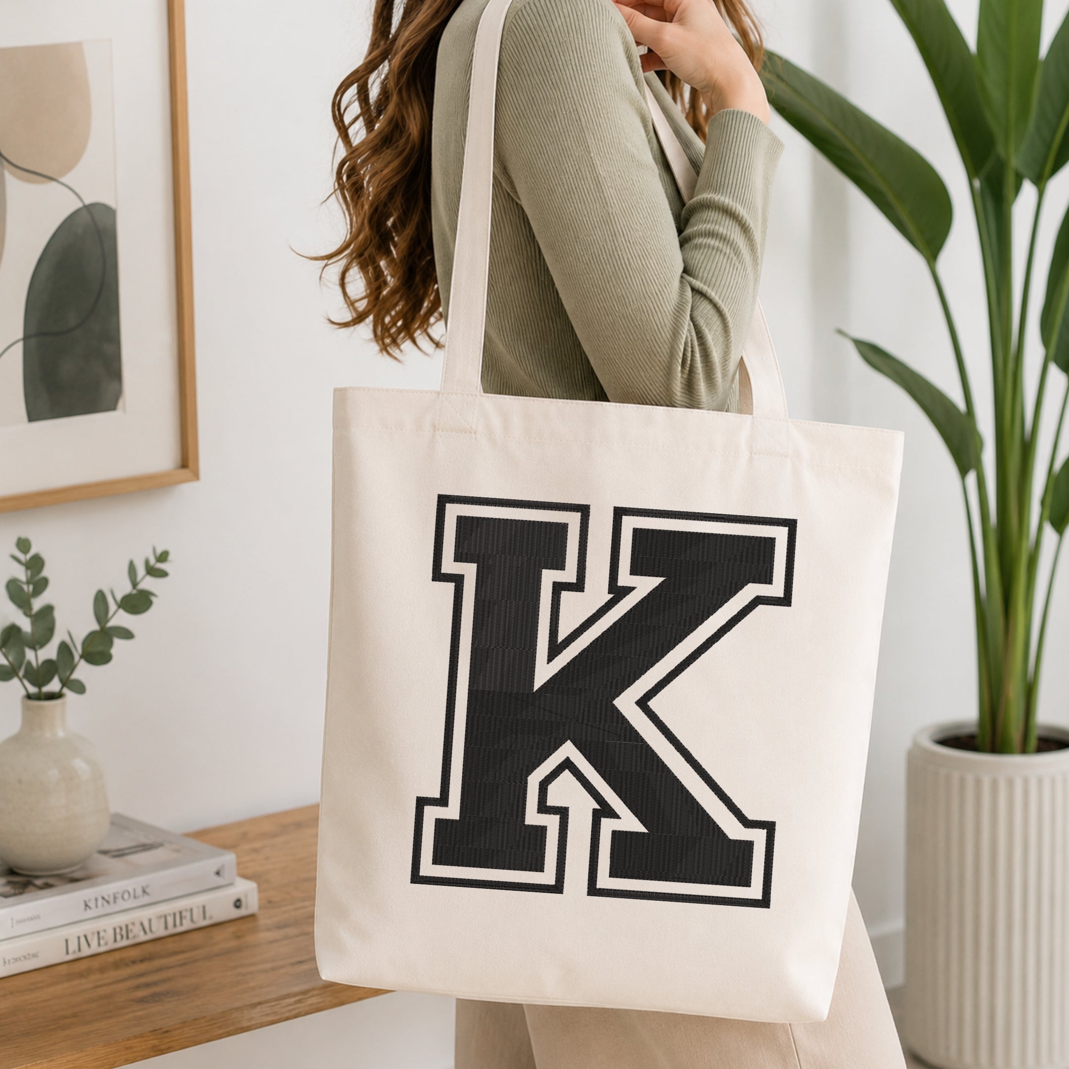 A woman wearing a sage green ribbed top carries a cream canvas tote bag with a large black block letter 'K' outlined in white on the front, set in a bright, modern room with a wooden console table and a potted plant.