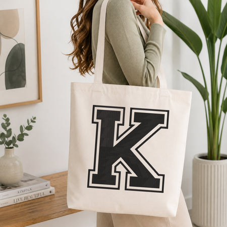 A woman wearing a sage green ribbed top carries a cream canvas tote bag with a large black block letter 'K' outlined in white on the front, set in a bright, modern room with a wooden console table and a potted plant.
