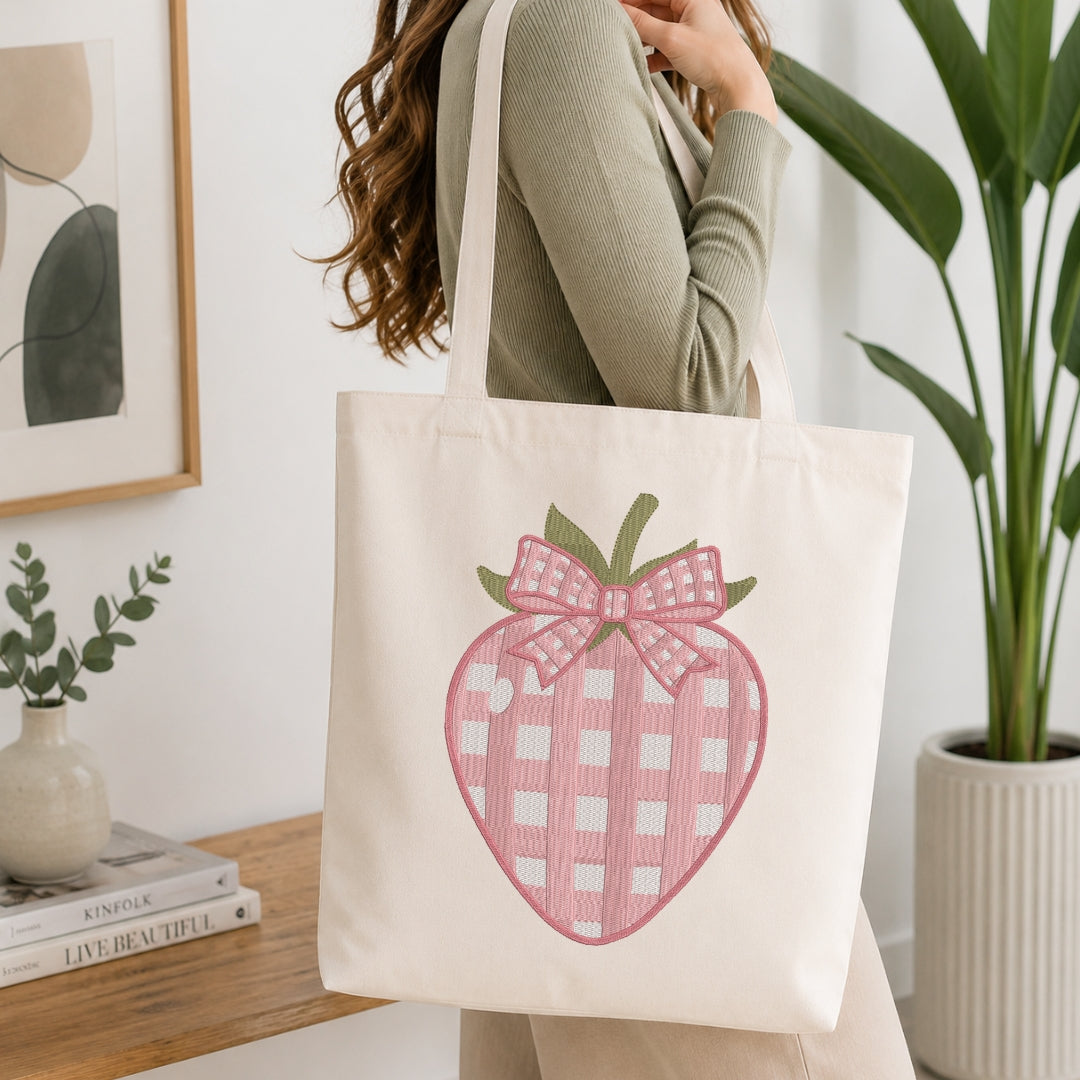 An off-white canvas tote bag with a pink and white gingham strawberry design and a matching bow, carried on the shoulder of a woman in a cozy indoor setting.