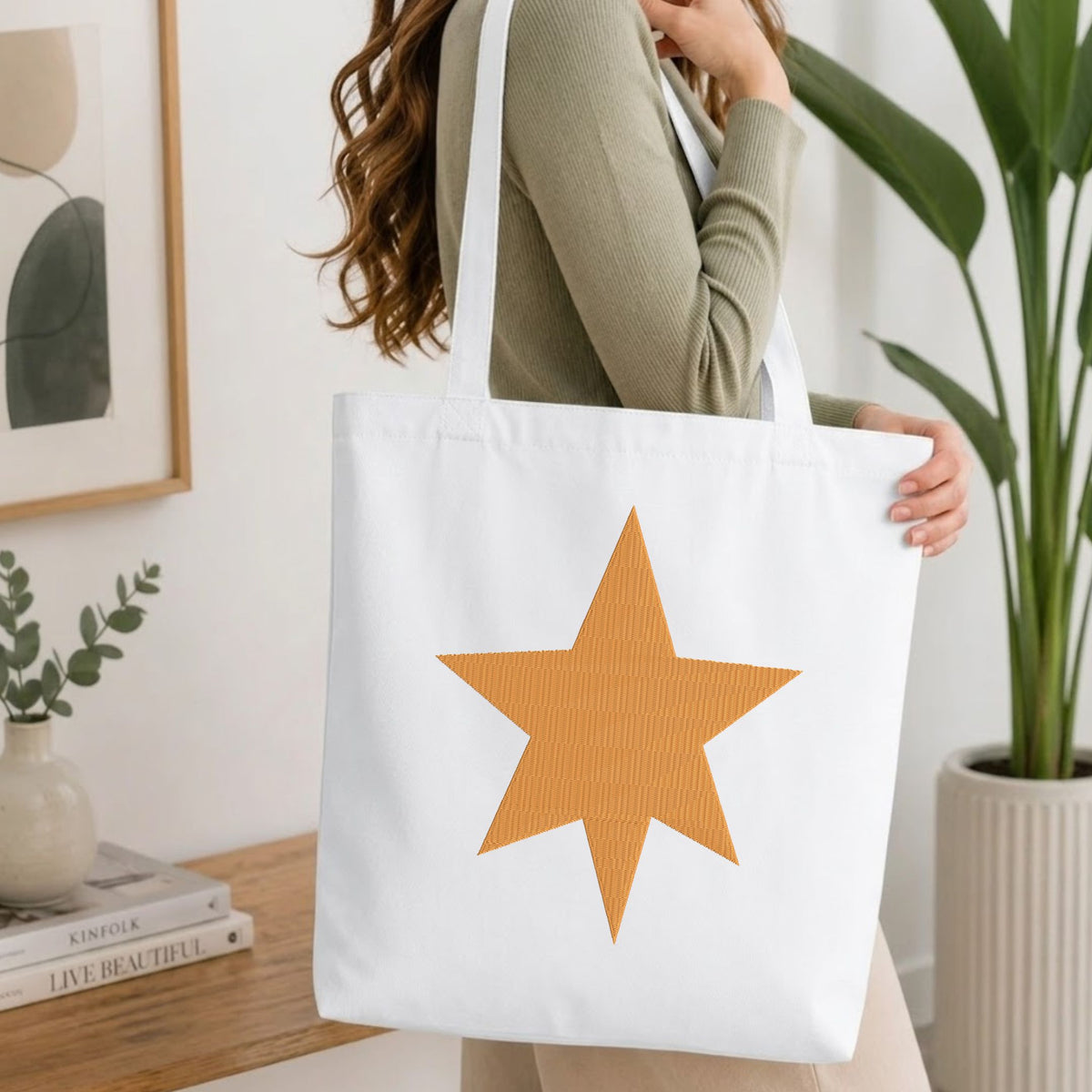 A woman wearing a sage green ribbed long-sleeve top carries a white canvas tote bag with a large textured orange star on the front, set in a bright, modern room with a wooden table, framed art, and a potted plant.